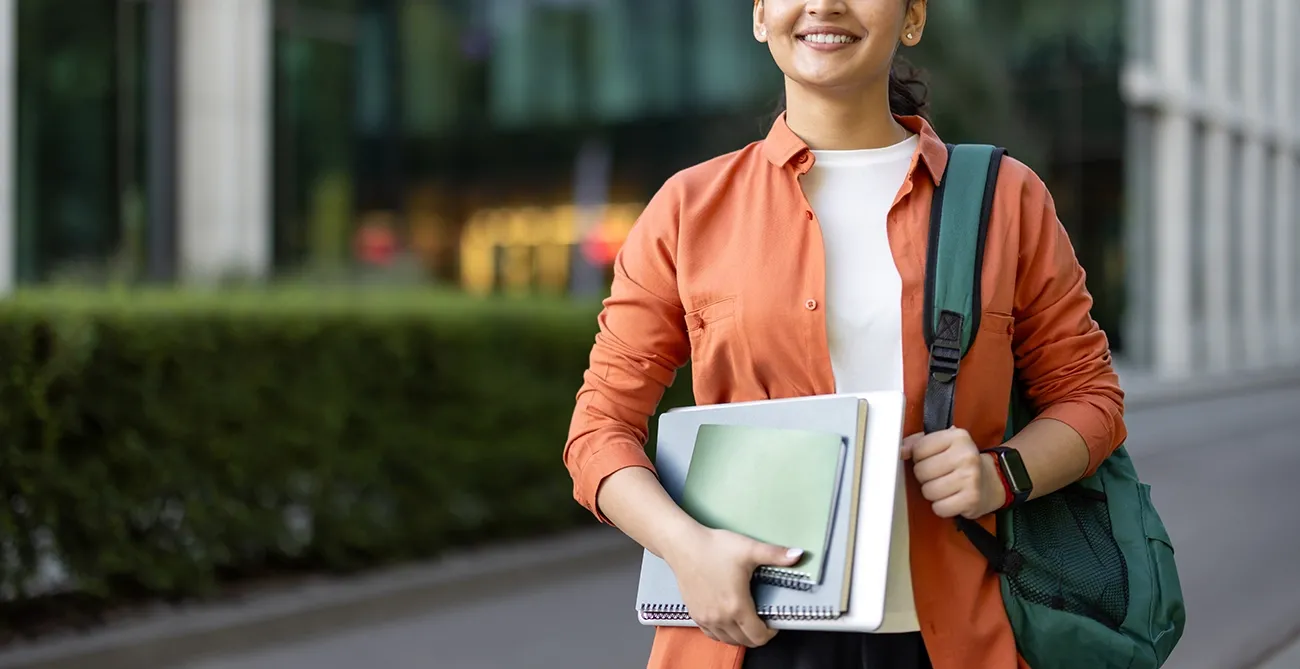 Female college student carrying a stack of notebooks with a backpack over one shoulder.