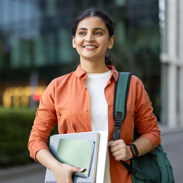 Female college student carrying a stack of notebooks with a backpack over one shoulder.