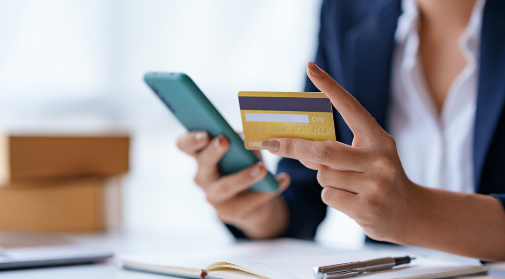 Businesswoman holds her smartphone in one hand and her debit card in another while sitting at a desk