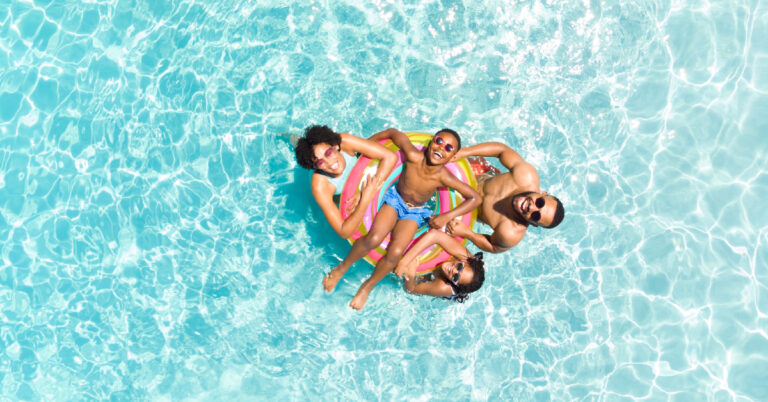 Black family of four spends time in the pool together. The son is floating in an innertube while the mom, dad, and siter surround him.