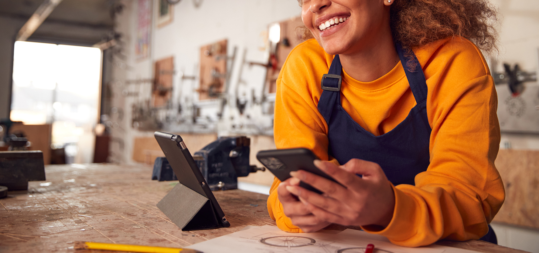Female Business Owner In Workshop Using Digital Tablet And Holding Mobile Phone