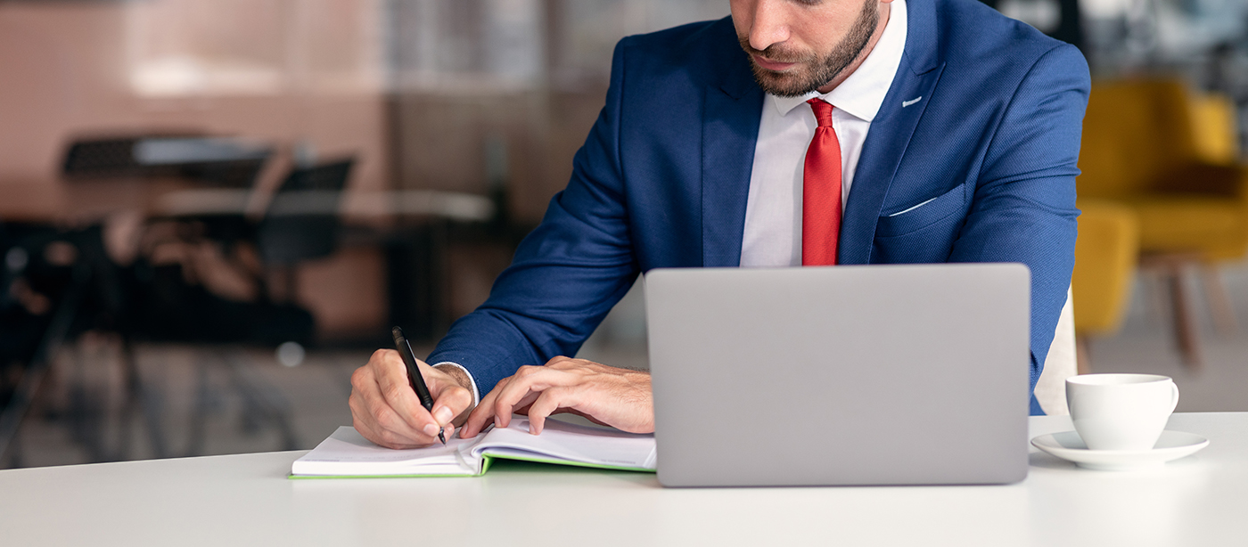 Businessman in a blue suit and red tie in a business office sitting at a desk. He is writing in a notebook with his laptop sitting on the desk in front of him.