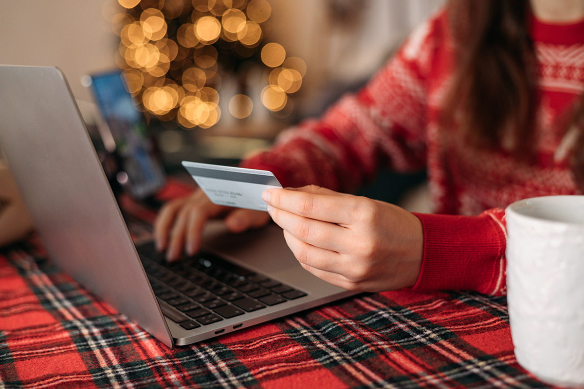 Hand with credit card on laptop with blank screen at table on background of stylish Christmas decorations, gifts and Christmas tree.