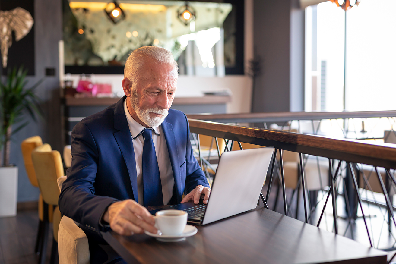 Older gentleman sitting in a coffee shop with a cup of coffee and his laptop.