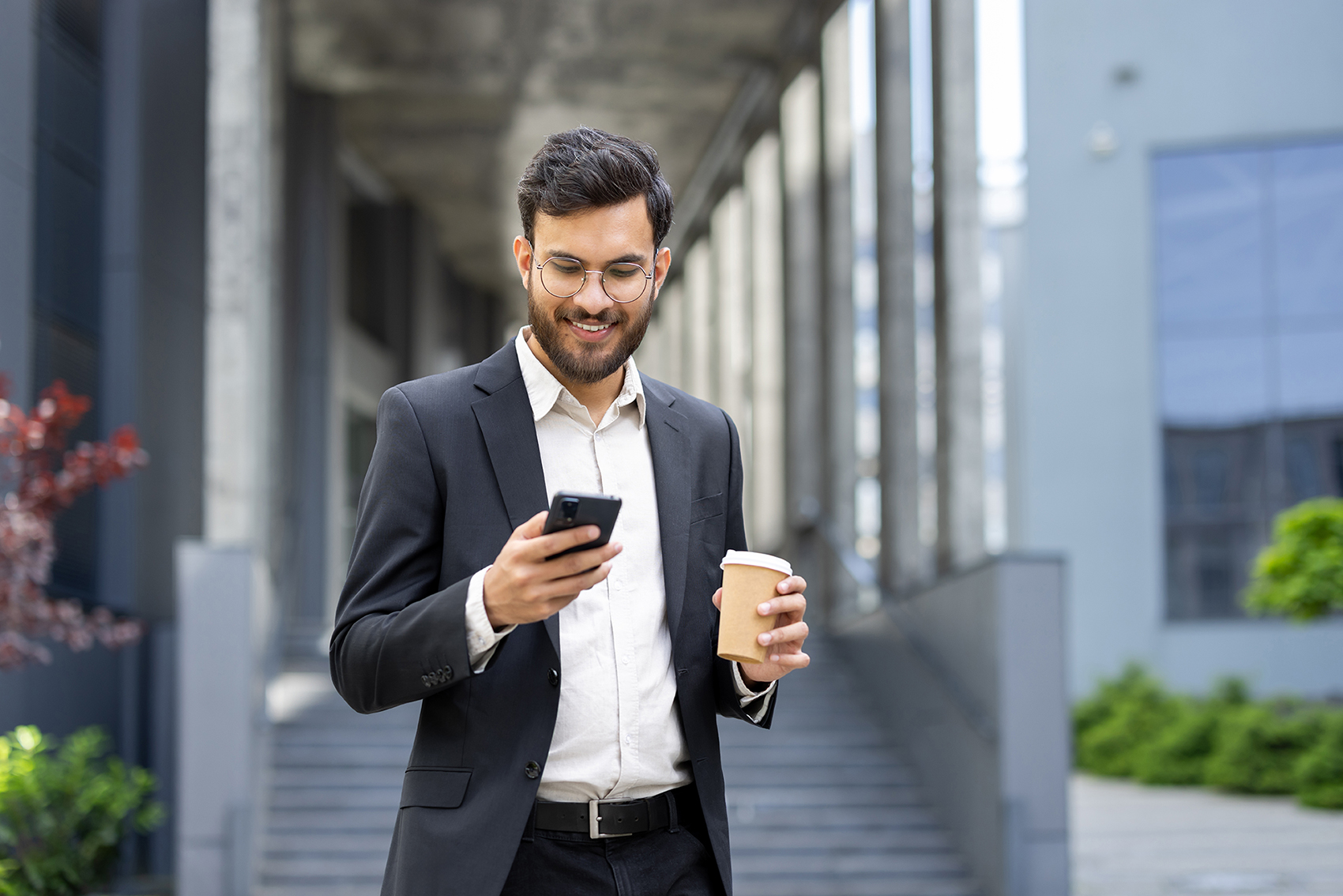 A smiling businessman in a suit checks his phone while holding a coffee cup outdoors.