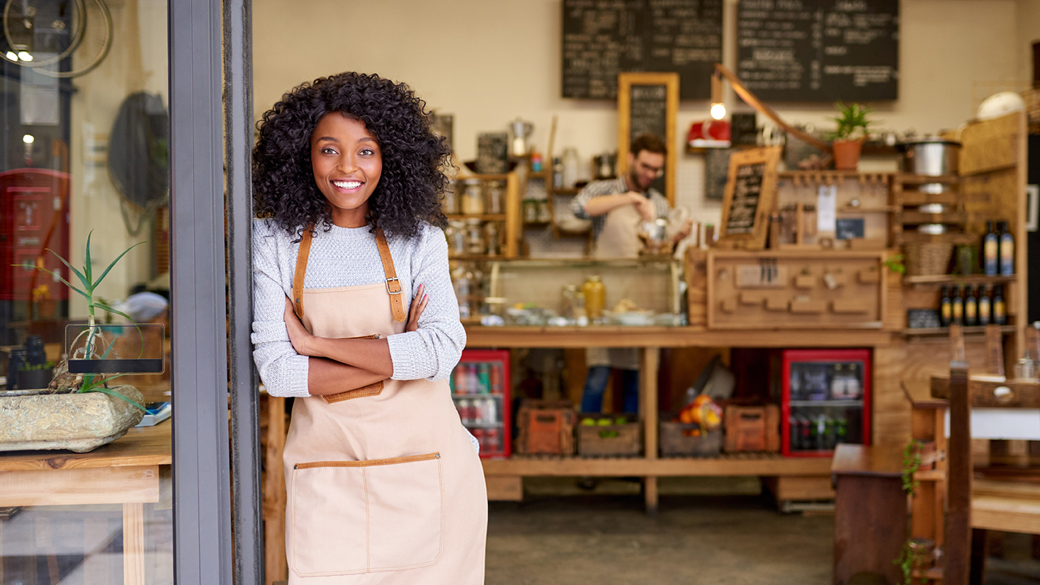 Portrait of a smiling young barista leaning with her arms crossed on the door of a trendy cafe