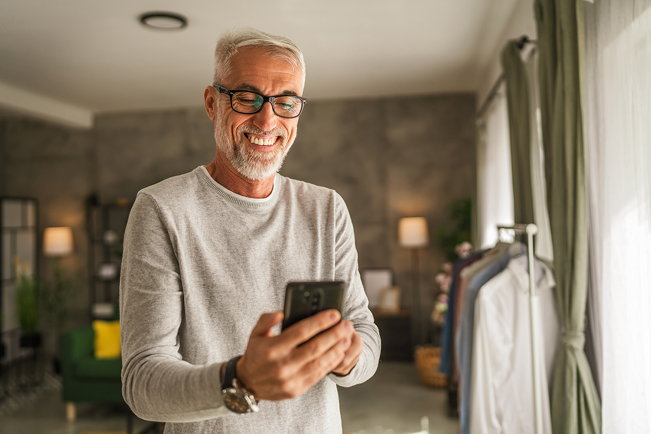 Mature caucasian man with eyeglasses hold mobile phone at home.
