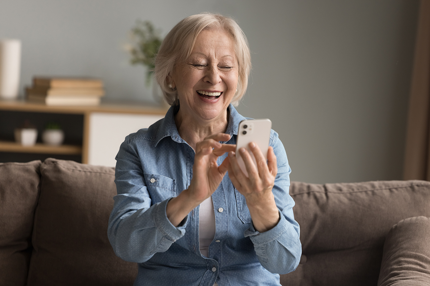 Happy grandmother sitting on sofa looking at phone screen with laughter.