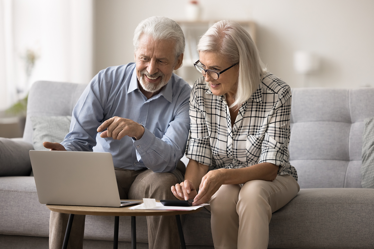 Smiling mature couple sit on sofa make budget analysis, using a calculator and laptop
