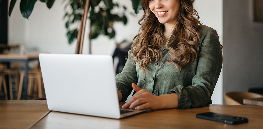 Smiling female using a laptop at a wooden table with a plant in the background.